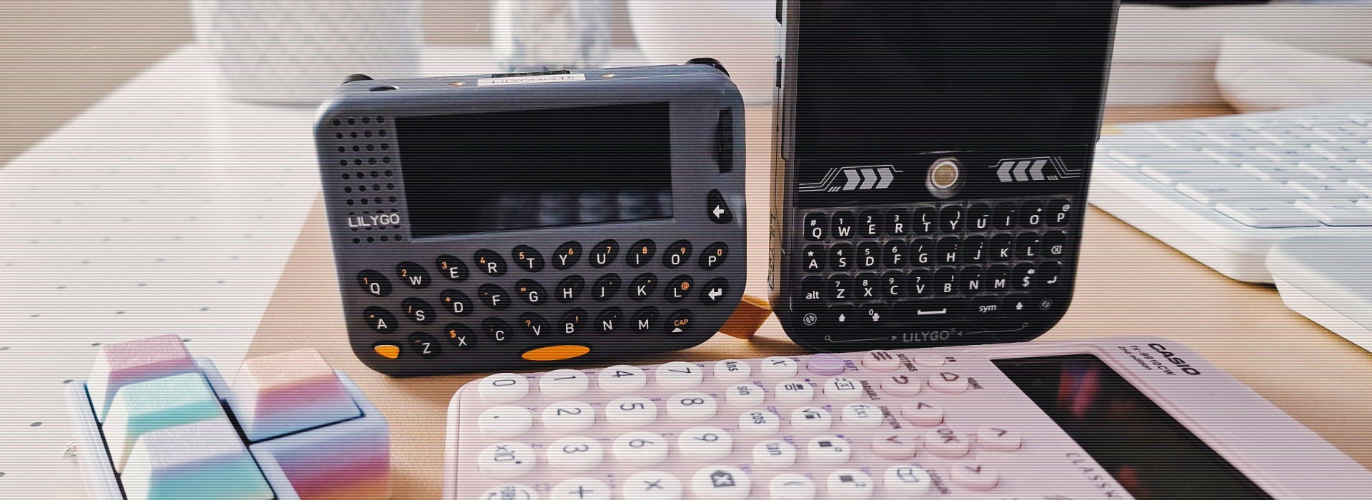 Image of the Lilygo T-Lora Pager and the Lilygo T-Deck side by side. In front of them, a pink Casio ClassWiz calulator.